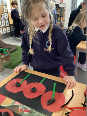 A young child in a school uniform standing over a table, engaged in a craft activity making three red poppies on a black background.