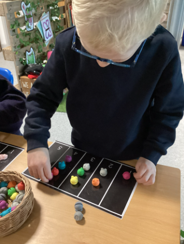 A young child wearing glasses and a uniform, sitting at a desk and sorting colorful, acorn-shaped counters onto a numbered black mat.