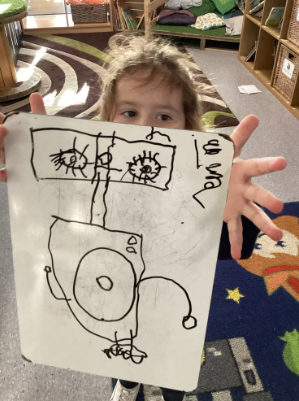A young child holding up a small whiteboard with a black marker drawing on it, partially obscuring their face.