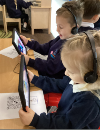 Two young children, wearing uniforms and headphones, sitting at a table and looking intently at tablets.