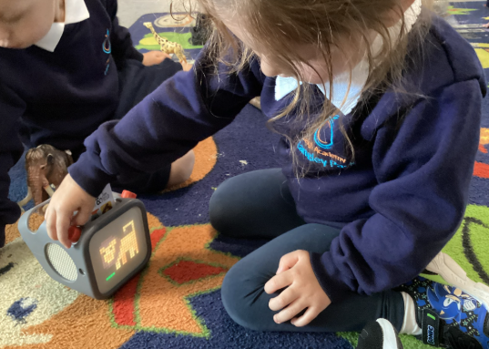 A young child sitting on the floor and interacting with a small, gray electronic device that displays a pixelated animal image.