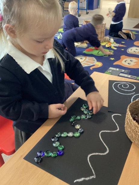 A young child placing green and clear glass beads along a curved line drawn with chalk on a black sheet of paper at a table.